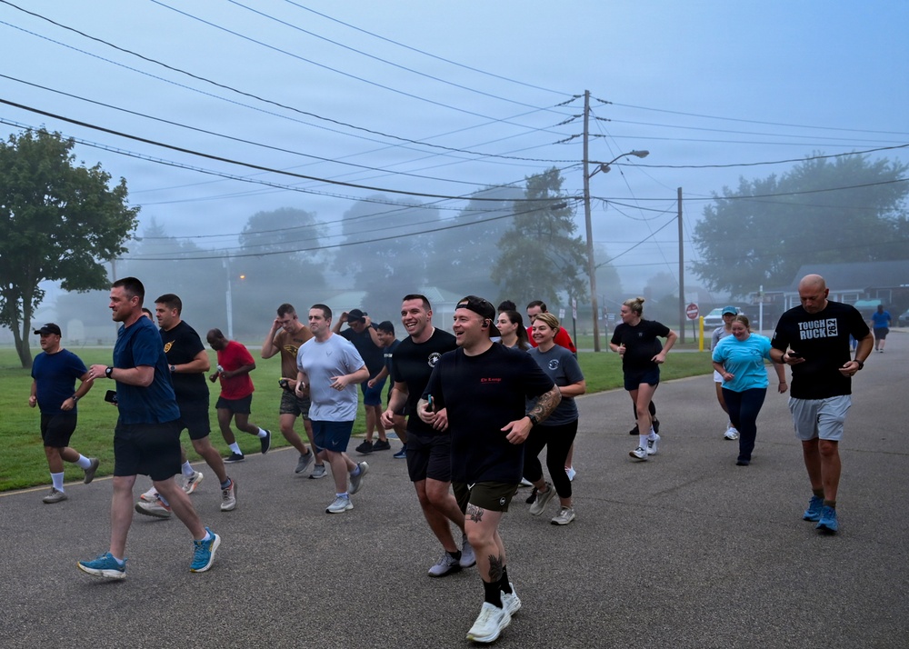 A large group of people are participating in an organized group run or fitness event on a paved road during early morning foggy conditions. Participants are dressed in casual athletic wear and appear to be in good spirits, with several individuals smiling and laughing. The setting appears to be a military base or institutional campus based on the uniform infrastructure and layout.