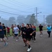 A large group of people are participating in an organized group run or fitness event on a paved road during early morning foggy conditions. Participants are dressed in casual athletic wear and appear to be in good spirits, with several individuals smiling and laughing. The setting appears to be a military base or institutional campus based on the uniform infrastructure and layout.