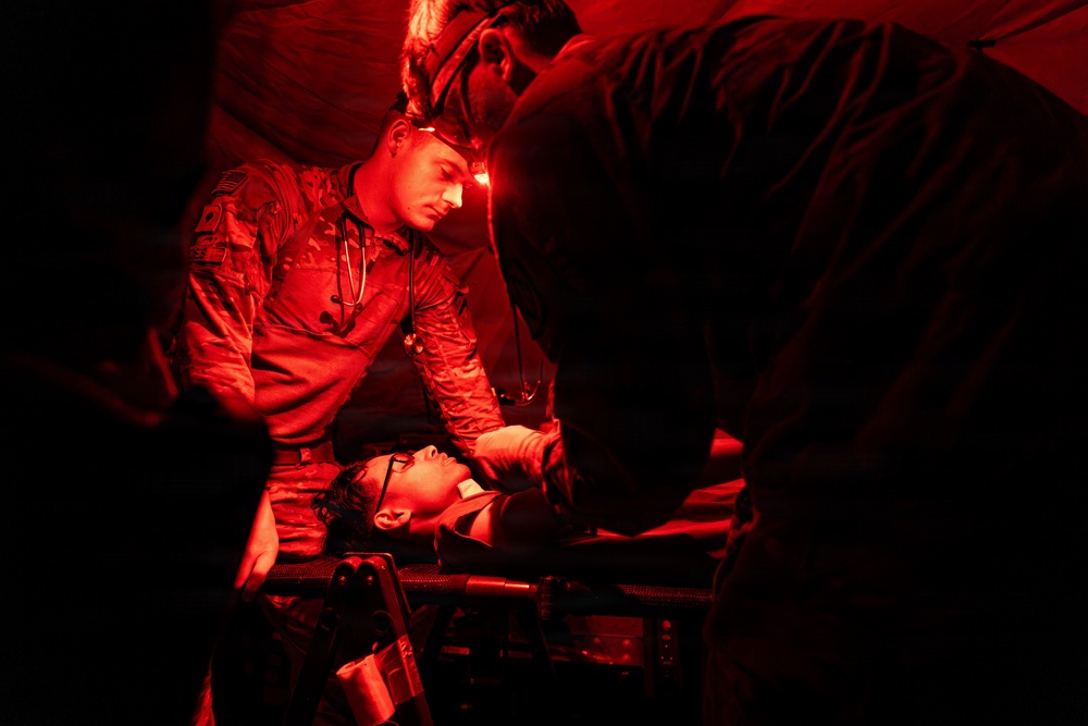 A military medic in camouflage uniform with a headlamp is treating a patient lying on a stretcher under red-light conditions, likely during a nighttime field medical exercise or operation. At least two personnel are attending to the patient in what appears to be a field medical tent or enclosed space. The red lighting is consistent with tactical night operations to preserve night vision.