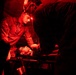 A military medic in camouflage uniform with a headlamp is treating a patient lying on a stretcher under red-light conditions, likely during a nighttime field medical exercise or operation. At least two personnel are attending to the patient in what appears to be a field medical tent or enclosed space. The red lighting is consistent with tactical night operations to preserve night vision.