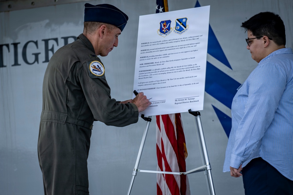 A U.S. Air Force officer in flight suit and service cap is signing an official proclamation document mounted on an easel, while a civilian woman observes. The document appears to be a Suicide Awareness and Prevention Month proclamation for Moody Air Force Base, dated September 1st. Two military unit insignia patches are visible at the top of the document along with an American flag in the background.