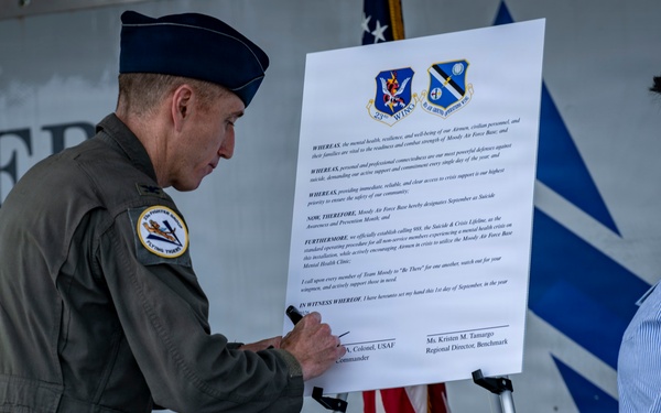 A U.S. Air Force officer in flight suit and service cap is signing an official proclamation document mounted on an easel, while a civilian woman observes. The document appears to be a Suicide Awareness and Prevention Month proclamation for Moody Air Force Base, dated September 1st. Two military unit insignia patches are visible at the top of the document along with an American flag in the background.