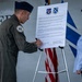 A U.S. Air Force officer in flight suit and service cap is signing an official proclamation document mounted on an easel, while a civilian woman observes. The document appears to be a Suicide Awareness and Prevention Month proclamation for Moody Air Force Base, dated September 1st. Two military unit insignia patches are visible at the top of the document along with an American flag in the background.