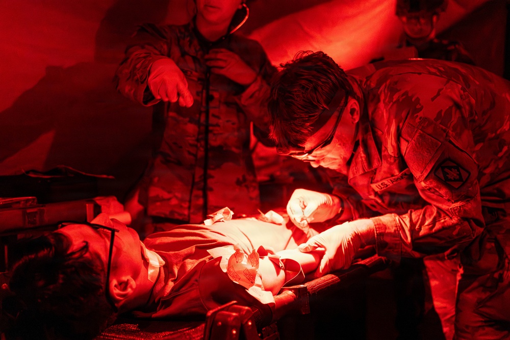 Military medical personnel in U.S. Army camouflage uniforms are performing a medical procedure or trauma care on a patient lying on a stretcher or field table. The scene is illuminated by red light, consistent with tactical/field medical operations where red lighting preserves night vision. At least three personnel are visible, with one actively treating the patient while wearing gloves.