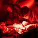 Military medical personnel in U.S. Army camouflage uniforms are performing a medical procedure or trauma care on a patient lying on a stretcher or field table. The scene is illuminated by red light, consistent with tactical/field medical operations where red lighting preserves night vision. At least three personnel are visible, with one actively treating the patient while wearing gloves.