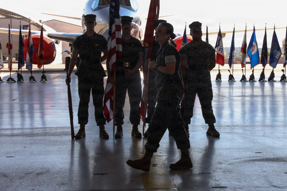 A military ceremony taking place inside an aircraft hangar, featuring personnel in camouflage uniforms participating in what appears to be a formal color guard or change of command ceremony. Multiple service members are holding flags including the American flag, with rows of unit/state guidon flags visible in the background. An aircraft is partially visible behind the formation.