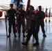 A military ceremony taking place inside an aircraft hangar, featuring personnel in camouflage uniforms participating in what appears to be a formal color guard or change of command ceremony. Multiple service members are holding flags including the American flag, with rows of unit/state guidon flags visible in the background. An aircraft is partially visible behind the formation.