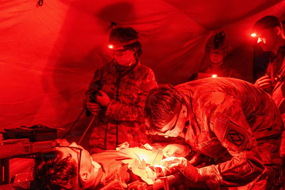 U.S. Army medical personnel are performing what appears to be an emergency field delivery or neonatal care inside a military field tent illuminated by red lighting. At least three soldiers in OCP (Operational Camouflage Pattern) uniforms are attending to a newborn infant on a makeshift surface. The scene suggests a forward operating medical unit conducting emergency obstetric or neonatal care under field conditions.