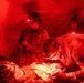 U.S. Army medical personnel are performing what appears to be an emergency field delivery or neonatal care inside a military field tent illuminated by red lighting. At least three soldiers in OCP (Operational Camouflage Pattern) uniforms are attending to a newborn infant on a makeshift surface. The scene suggests a forward operating medical unit conducting emergency obstetric or neonatal care under field conditions.