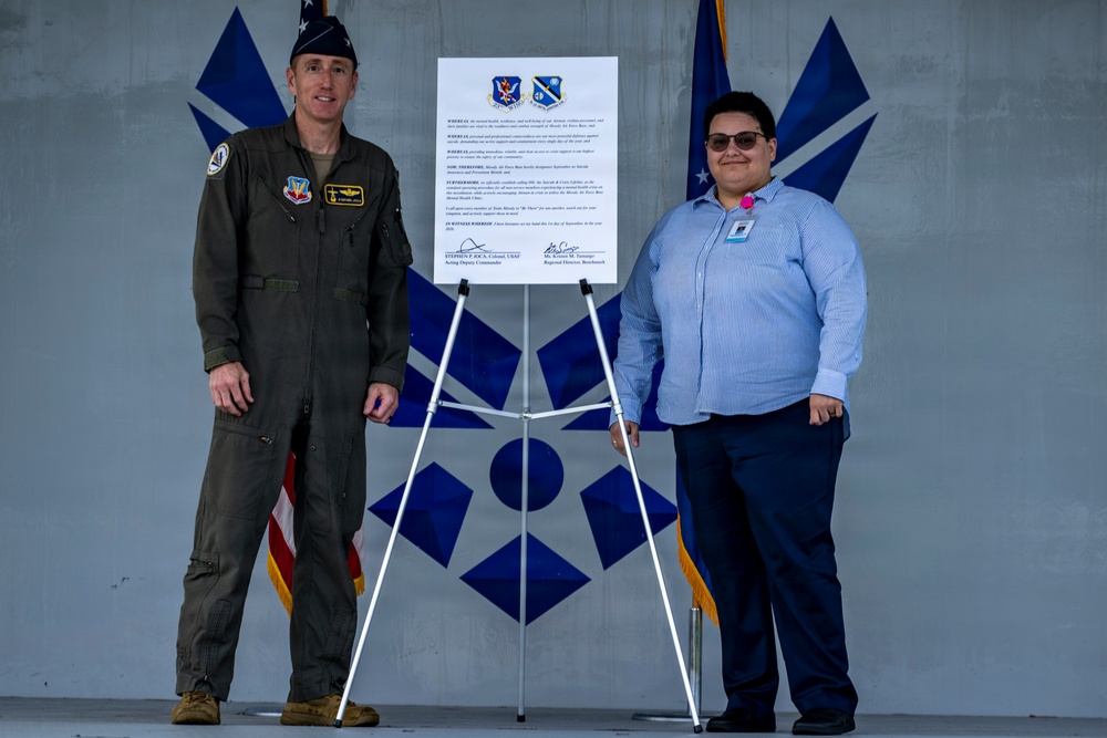 Two individuals stand on either side of a mounted proclamation or official document on an easel, posing for a formal photograph. The setting appears to be a military installation, with U.S. Air Force flags and insignia visible in the background. One individual is in a military flight suit and the other is in civilian business attire.