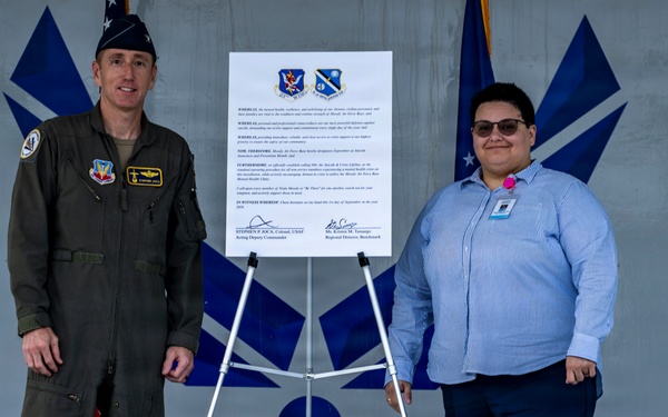 Two individuals stand on either side of a mounted proclamation or official document on an easel, posing for a formal photograph. The setting appears to be a military installation, with U.S. Air Force flags and insignia visible in the background. One individual is in a military flight suit and the other is in civilian business attire.