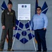 Two individuals stand on either side of a mounted proclamation or official document on an easel, posing for a formal photograph. The setting appears to be a military installation, with U.S. Air Force flags and insignia visible in the background. One individual is in a military flight suit and the other is in civilian business attire.