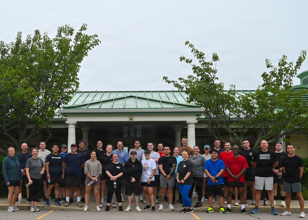 A large group of approximately 35-40 people posed for a group photo in a parking lot in front of a building with a green metal roof and white columns. The participants are dressed in casual athletic/workout attire, suggesting this is a fitness or team-building event. The overcast sky and lush green trees frame the scene.