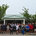 A large group of approximately 35-40 people posed for a group photo in a parking lot in front of a building with a green metal roof and white columns. The participants are dressed in casual athletic/workout attire, suggesting this is a fitness or team-building event. The overcast sky and lush green trees frame the scene.