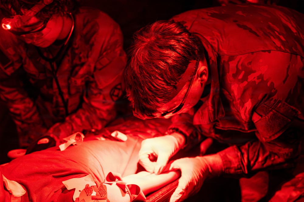 Two individuals in military camouflage uniforms are performing what appears to be a medical procedure or first aid treatment on a patient under red-tinted lighting conditions. One person is wearing white latex gloves and is actively working on the patient's arm or hand area. The scene is consistent with a tactical combat casualty care (TCCC) training exercise or field medical treatment scenario.