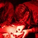 Two individuals in military camouflage uniforms are performing what appears to be a medical procedure or first aid treatment on a patient under red-tinted lighting conditions. One person is wearing white latex gloves and is actively working on the patient's arm or hand area. The scene is consistent with a tactical combat casualty care (TCCC) training exercise or field medical treatment scenario.