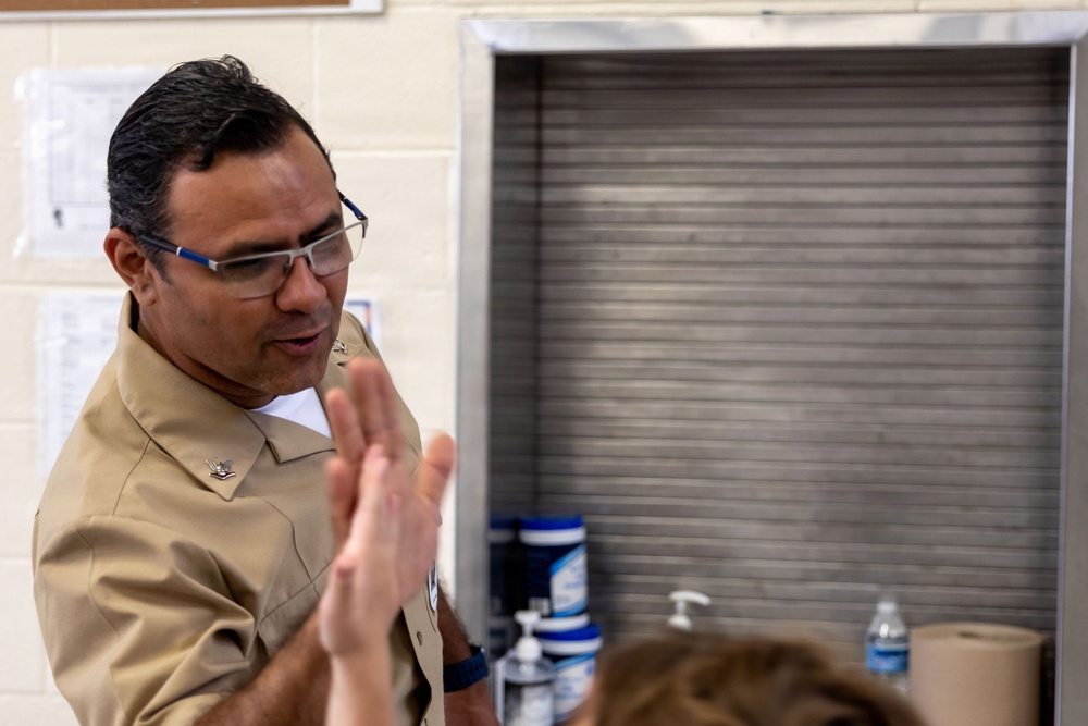 A man wearing a tan/khaki military-style uniform with a collar insignia is engaged in conversation or instruction, gesturing with his hands. He is wearing rectangular glasses with blue accents and appears to be speaking to someone partially visible in the foreground. The setting appears to be an indoor facility with a cinder block wall and a metal roll-up door or cabinet in the background.