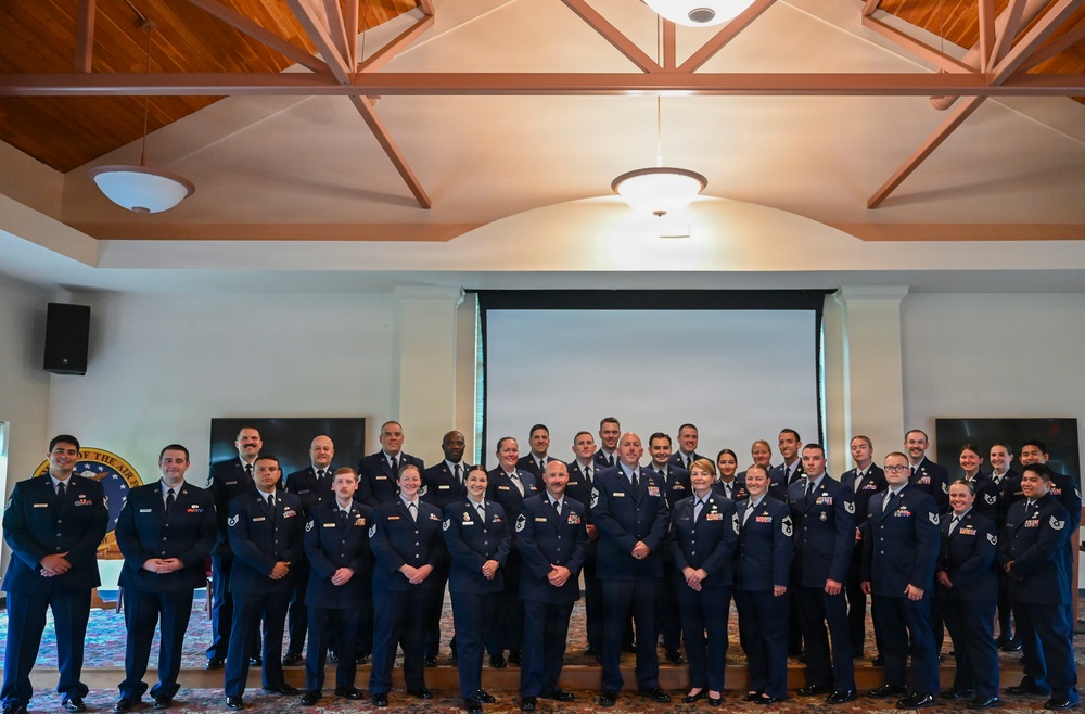A large group of United States Air Force personnel in dress blue uniforms are posed for a formal group photograph inside what appears to be a banquet or event hall. The group consists of approximately 30 individuals arranged in two to three rows, including both male and female service members of various ranks. A projection screen, wooden-beamed ceiling, and a partial Air Force seal are visible in the background.