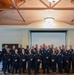 A large group of United States Air Force personnel in dress blue uniforms are posed for a formal group photograph inside what appears to be a banquet or event hall. The group consists of approximately 30 individuals arranged in two to three rows, including both male and female service members of various ranks. A projection screen, wooden-beamed ceiling, and a partial Air Force seal are visible in the background.