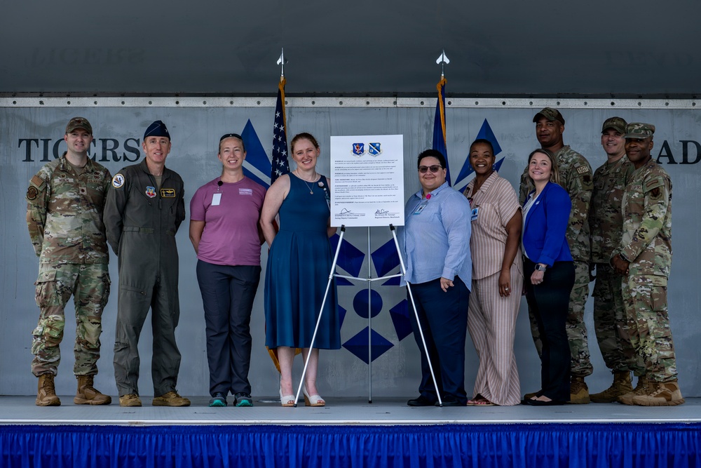 A group of approximately ten individuals, including military personnel in uniform and civilians in business casual attire, pose together on a stage in front of a document displayed on an easel. The setting appears to be a military installation hangar or facility, with partial text reading 'TIGERS' and 'AD' visible on the backdrop. The occasion appears to be a formal signing ceremony or official agreement, indicated by the framed document bearing two military insignia patches.