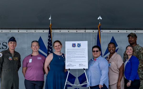 A group of approximately ten individuals, including military personnel in uniform and civilians in business casual attire, pose together on a stage in front of a document displayed on an easel. The setting appears to be a military installation hangar or facility, with partial text reading 'TIGERS' and 'AD' visible on the backdrop. The occasion appears to be a formal signing ceremony or official agreement, indicated by the framed document bearing two military insignia patches.