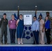 A group of approximately ten individuals, including military personnel in uniform and civilians in business casual attire, pose together on a stage in front of a document displayed on an easel. The setting appears to be a military installation hangar or facility, with partial text reading 'TIGERS' and 'AD' visible on the backdrop. The occasion appears to be a formal signing ceremony or official agreement, indicated by the framed document bearing two military insignia patches.