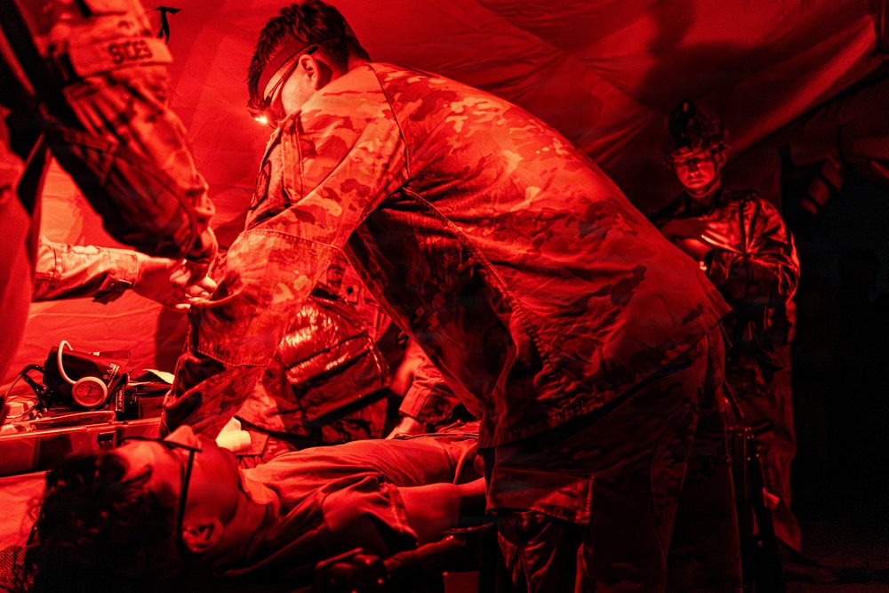 Military personnel are conducting a medical treatment or triage procedure inside a field tent illuminated by red lighting. A soldier in camouflage uniform leans over a patient lying on a surface, performing what appears to be a medical assessment or intervention. At least two other personnel are present in the background observing or assisting.