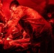 Military personnel are conducting a medical treatment or triage procedure inside a field tent illuminated by red lighting. A soldier in camouflage uniform leans over a patient lying on a surface, performing what appears to be a medical assessment or intervention. At least two other personnel are present in the background observing or assisting.
