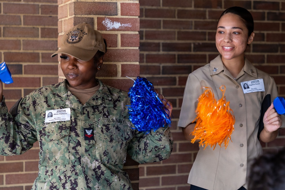 Two U.S. Navy personnel in uniform are holding cheerleader pom-poms — one blue and one orange — appearing to participate in a morale or community outreach event. Both individuals are wearing visitor badges and are standing near a brick wall. The person on the left wears digital camouflage NWU (Navy Working Uniform) with a U.S.S. Wasp cap, while the person on the right wears a khaki service uniform.