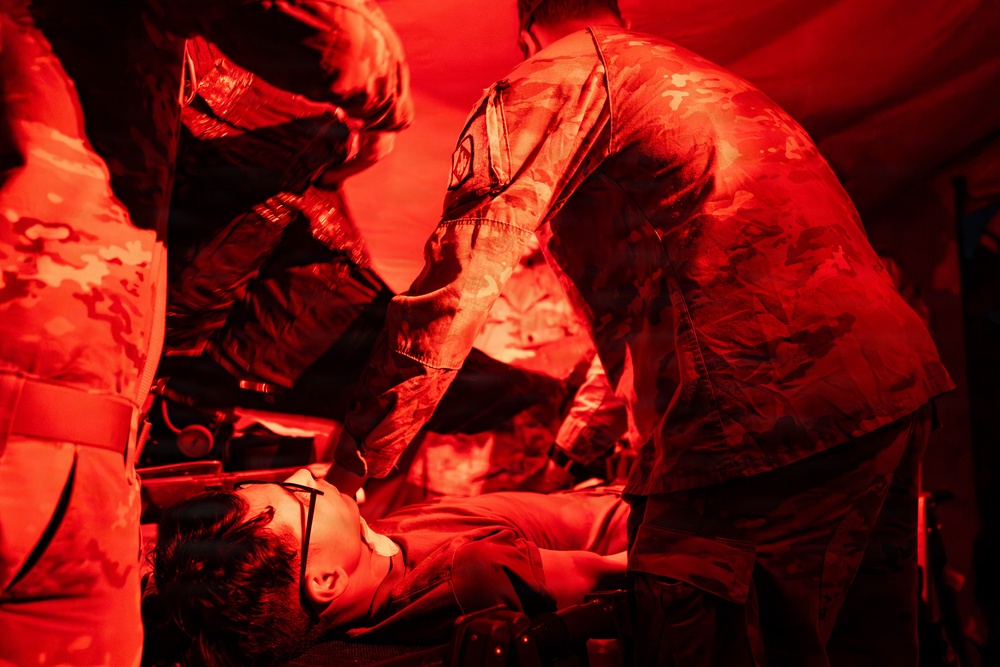 Military medical personnel in camouflage uniforms are attending to a patient lying on a stretcher or litter under intense red lighting, consistent with a field medical treatment scenario. The red lighting suggests a tactical or low-light medical environment, possibly a field hospital or medical training exercise. At least two uniformed individuals are actively working on the patient.