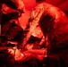 Military medical personnel in camouflage uniforms are attending to a patient lying on a stretcher or litter under intense red lighting, consistent with a field medical treatment scenario. The red lighting suggests a tactical or low-light medical environment, possibly a field hospital or medical training exercise. At least two uniformed individuals are actively working on the patient.