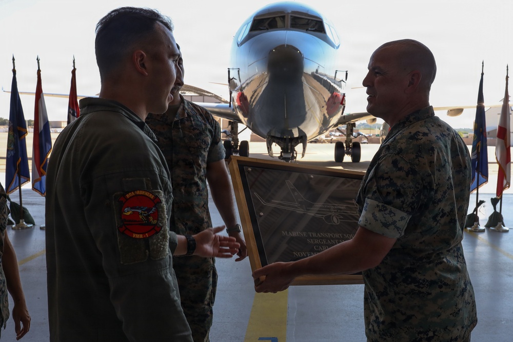 A military ceremony taking place in an aircraft hangar or tarmac area, where U.S. Marine Corps personnel are presenting a framed commemorative plaque or award. Three Marines in camouflage uniforms are visible, with one wearing a flight suit bearing a squadron patch. A military aircraft is visible in the background along with multiple unit or state flags.