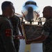 A military ceremony taking place in an aircraft hangar or tarmac area, where U.S. Marine Corps personnel are presenting a framed commemorative plaque or award. Three Marines in camouflage uniforms are visible, with one wearing a flight suit bearing a squadron patch. A military aircraft is visible in the background along with multiple unit or state flags.