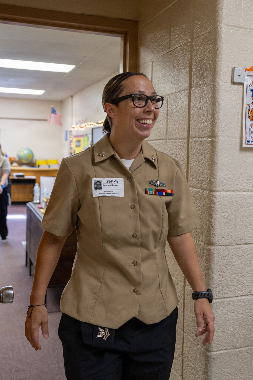A female U.S. Navy service member in tan Service Uniform (NWU Type III or Service Khaki) is walking through a doorway in what appears to be an elementary school hallway. She is smiling and wearing a visitor badge identifying her as 'Keresea Illenye' from 'Tarrallton Elementary School.' She displays several service ribbons, a warfare device, and collar insignia consistent with a senior enlisted or officer rank.