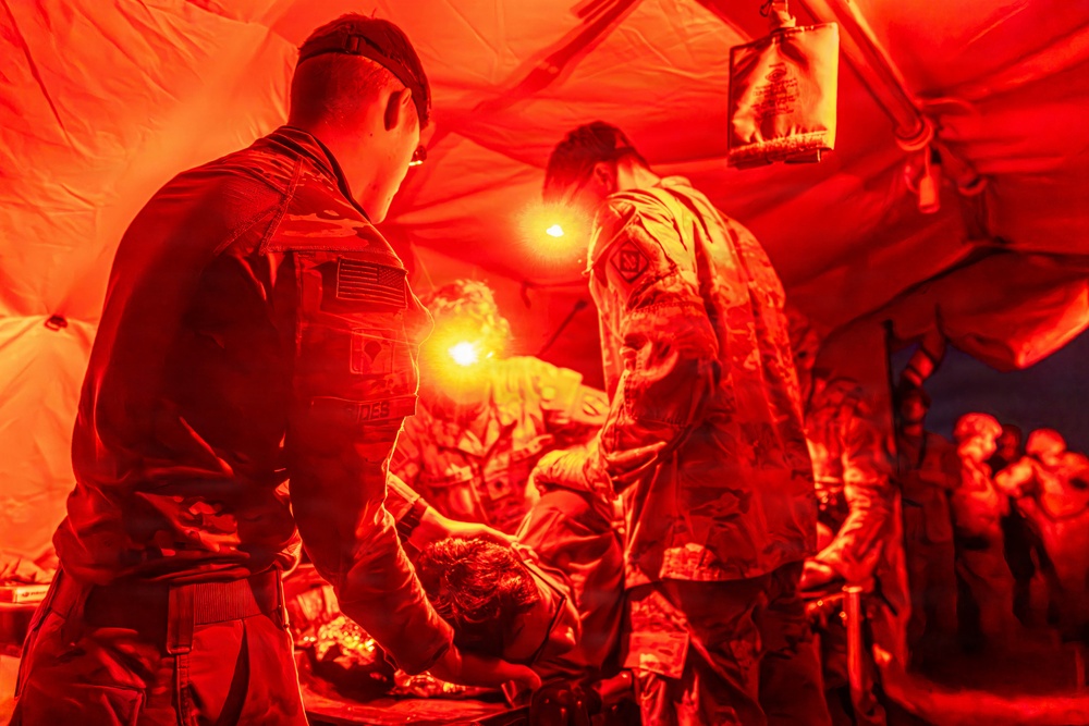 Military medical personnel are treating a patient inside a field medical tent illuminated by red lighting. At least two uniformed soldiers wearing U.S. Army patches are attending to a person lying on what appears to be a stretcher or treatment surface. The scene appears to depict a combat casualty care or field triage exercise/operation.