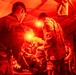 Military medical personnel are treating a patient inside a field medical tent illuminated by red lighting. At least two uniformed soldiers wearing U.S. Army patches are attending to a person lying on what appears to be a stretcher or treatment surface. The scene appears to depict a combat casualty care or field triage exercise/operation.
