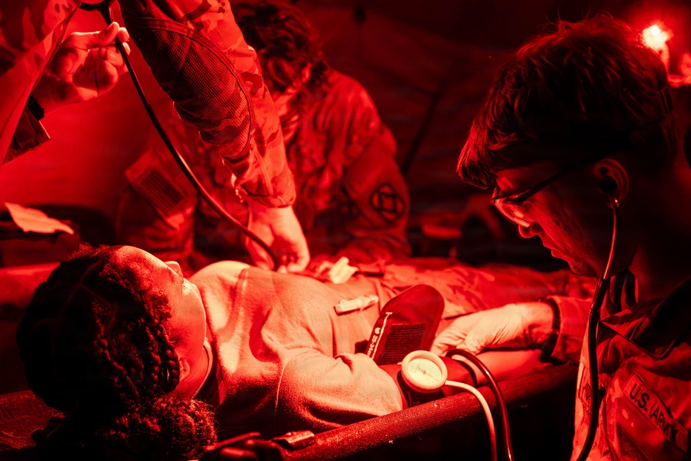 U.S. Army medical personnel are treating a patient on a stretcher under red-light conditions, consistent with a field medical or combat support hospital environment. At least two or three personnel in military uniforms are performing medical assessments including blood pressure measurement and auscultation with a stethoscope. The red lighting suggests a tactical or low-light operational medical setting.