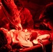 U.S. Army medical personnel are treating a patient on a stretcher under red-light conditions, consistent with a field medical or combat support hospital environment. At least two or three personnel in military uniforms are performing medical assessments including blood pressure measurement and auscultation with a stethoscope. The red lighting suggests a tactical or low-light operational medical setting.