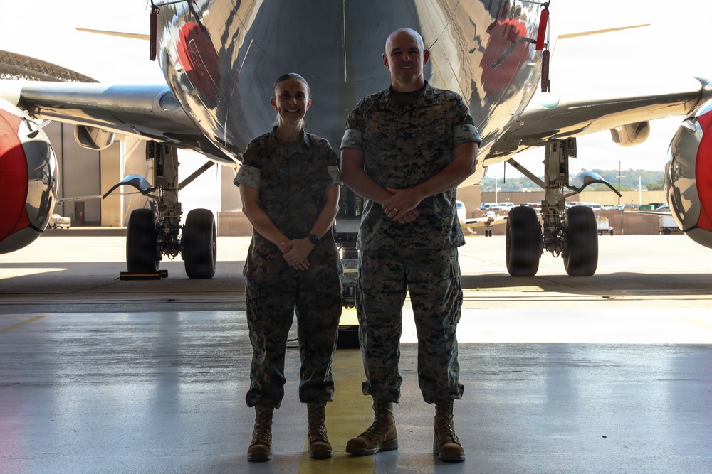 Two military personnel in matching camouflage uniforms stand side by side beneath the fuselage of a large aircraft inside a hangar. Both individuals are smiling and standing at ease with hands clasped in front of them. The setting appears to be a military aviation facility with the aircraft's landing gear and red-capped engine nacelles visible in the background.