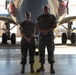 Two military personnel in matching camouflage uniforms stand side by side beneath the fuselage of a large aircraft inside a hangar. Both individuals are smiling and standing at ease with hands clasped in front of them. The setting appears to be a military aviation facility with the aircraft's landing gear and red-capped engine nacelles visible in the background.