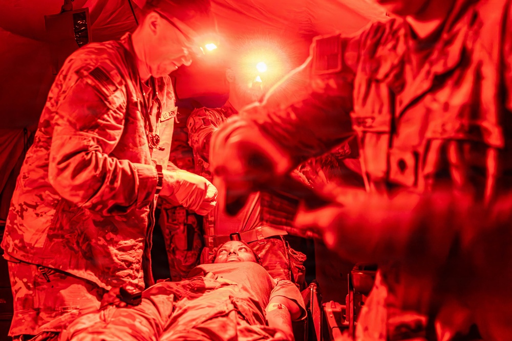 Military medical personnel are treating a casualty on a stretcher inside what appears to be a field medical tent illuminated by red lighting. At least two uniformed soldiers wearing camouflage uniforms are attending to a patient lying on a litter. The scene suggests a combat casualty care or tactical combat casualty care (TCCC) scenario, possibly a training exercise or real field medical operation.