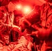 Military medical personnel are treating a casualty on a stretcher inside what appears to be a field medical tent illuminated by red lighting. At least two uniformed soldiers wearing camouflage uniforms are attending to a patient lying on a litter. The scene suggests a combat casualty care or tactical combat casualty care (TCCC) scenario, possibly a training exercise or real field medical operation.