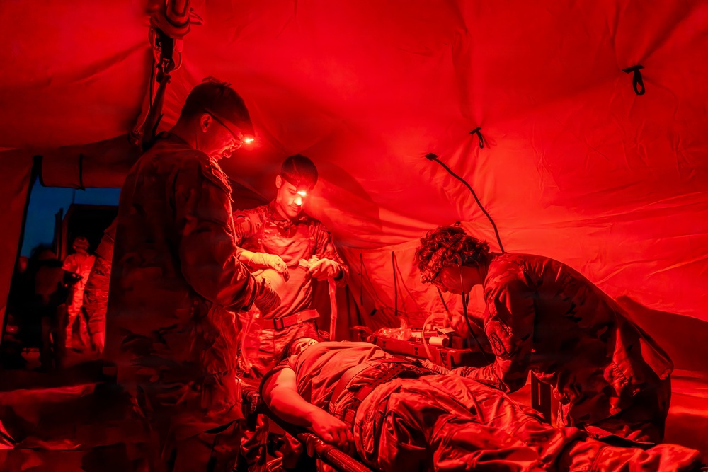 Military medical personnel are treating a patient on a field stretcher inside a tactical tent illuminated by red lighting. Three uniformed individuals wearing headlamps are actively performing medical procedures on a supine patient. The scene appears to be a combat casualty care or field medical training exercise conducted at night.