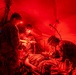 Military medical personnel are treating a patient on a field stretcher inside a tactical tent illuminated by red lighting. Three uniformed individuals wearing headlamps are actively performing medical procedures on a supine patient. The scene appears to be a combat casualty care or field medical training exercise conducted at night.