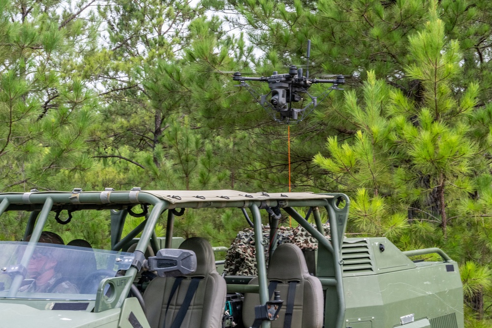 A military-style operation is depicted with a tethered drone hovering above a green tactical utility vehicle in a forested area. The drone appears to be connected to the vehicle via an orange tether cable, suggesting a tethered UAS (Unmanned Aerial System) configuration for persistent surveillance. The setting appears to be a training or demonstration exercise in a pine forest environment.