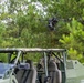 A military-style operation is depicted with a tethered drone hovering above a green tactical utility vehicle in a forested area. The drone appears to be connected to the vehicle via an orange tether cable, suggesting a tethered UAS (Unmanned Aerial System) configuration for persistent surveillance. The setting appears to be a training or demonstration exercise in a pine forest environment.