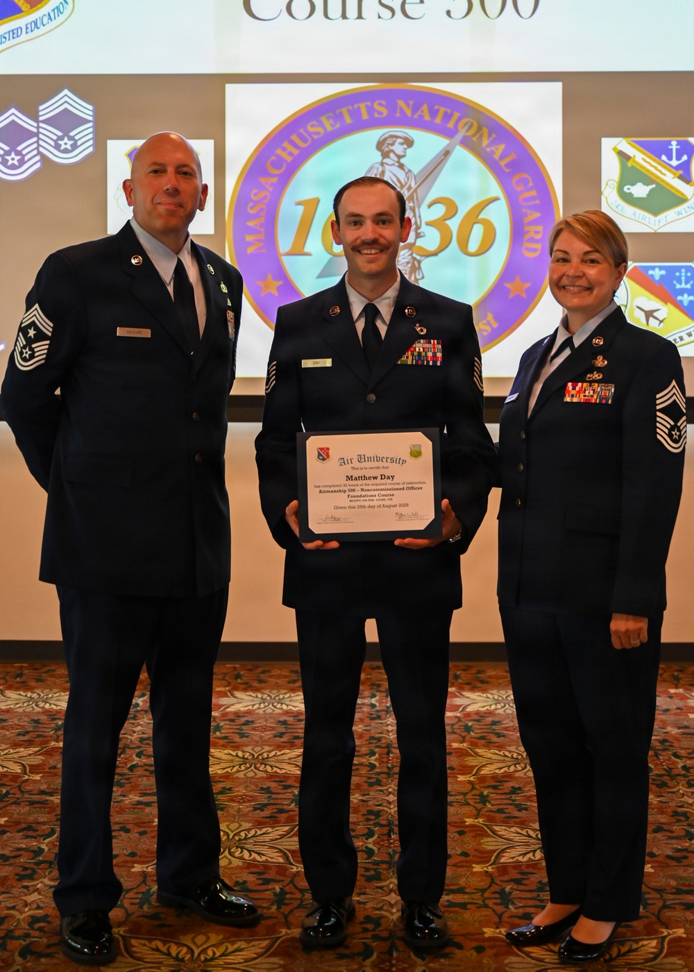 Three U.S. Air Force personnel in dress blue uniforms pose for a formal photo at what appears to be a graduation or awards ceremony. The central figure, identified as Matthew Day on his name tag, holds an Air University certificate for completing the Airmanship 500 - Noncommissioned Officers Foundations Course. The event is associated with the Massachusetts National Guard, as indicated by the projected logo in the background.