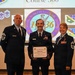 Three U.S. Air Force personnel in dress blue uniforms pose for a formal photo at what appears to be a graduation or awards ceremony. The central figure, identified as Matthew Day on his name tag, holds an Air University certificate for completing the Airmanship 500 - Noncommissioned Officers Foundations Course. The event is associated with the Massachusetts National Guard, as indicated by the projected logo in the background.