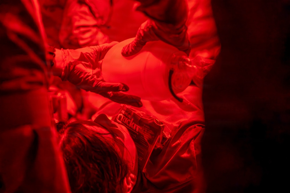 The image is bathed entirely in intense red lighting, making details difficult to discern. Multiple individuals appear to be wearing protective or specialized suits, with one person handling a smooth, rounded white object. A partially visible label or packaging with text reading 'Quick' or similar is visible on one of the suits.