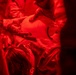 The image is bathed entirely in intense red lighting, making details difficult to discern. Multiple individuals appear to be wearing protective or specialized suits, with one person handling a smooth, rounded white object. A partially visible label or packaging with text reading 'Quick' or similar is visible on one of the suits.